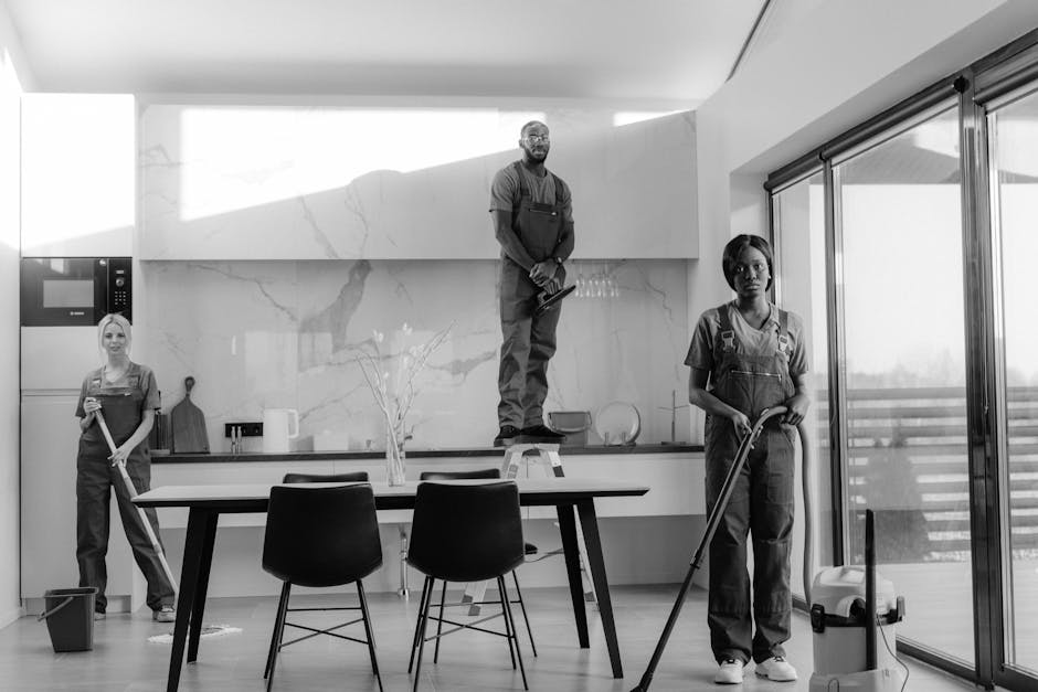 A black-and-white photograph of three professional cleaners in a modern kitchen, each wearing uniform overalls. One cleaner is standing on a step ladder, holding a cleaning cloth, while another stands near the window holding a mop. The third cleaner is at the side, also with a mop, near a small bucket on the floor. The kitchen features a large, polished marble backsplash, a sleek white countertop, and a glass door leading to an outdoor balcony. The space appears highly clean, with surfaces well-maintained and reflecting light, emphasizing thorough surface cleaning and sanitisation by Knightsbridge Cleaner in a domestic setting.