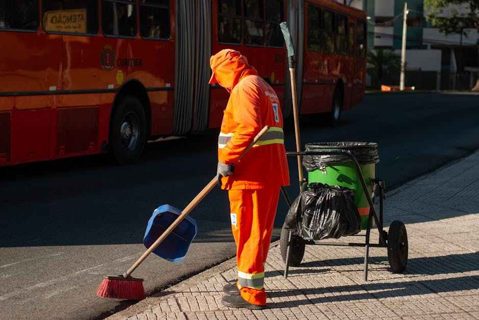 A street cleaner dressed in bright orange high-visibility clothing, including a jacket and pants, is sweeping the pavement with a long-handled broom. The cleaner's head is covered with an orange hood or cap, and they are wearing dark gloves. Next to them is a green waste collection bin on a wheeled cart lined with a black trash bag, with additional cleaning tools attached. In the background, there is a red bus parked on the street, with street and building details visible in daylight, indicating an urban environment. The overall scene depicts thorough surface cleaning and street maintenance, representative of professional cleaning services, with Knightsbridge Cleaner delivering local sanitation and deep cleaning solutions.