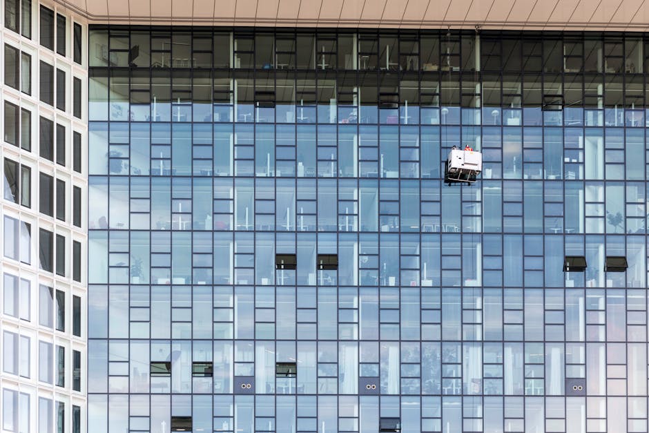 A high-rise building with a glass facade featuring multiple reflective windows arranged in a grid pattern. Several window cleaners wearing yellow safety harnesses and helmets are suspended from ropes,