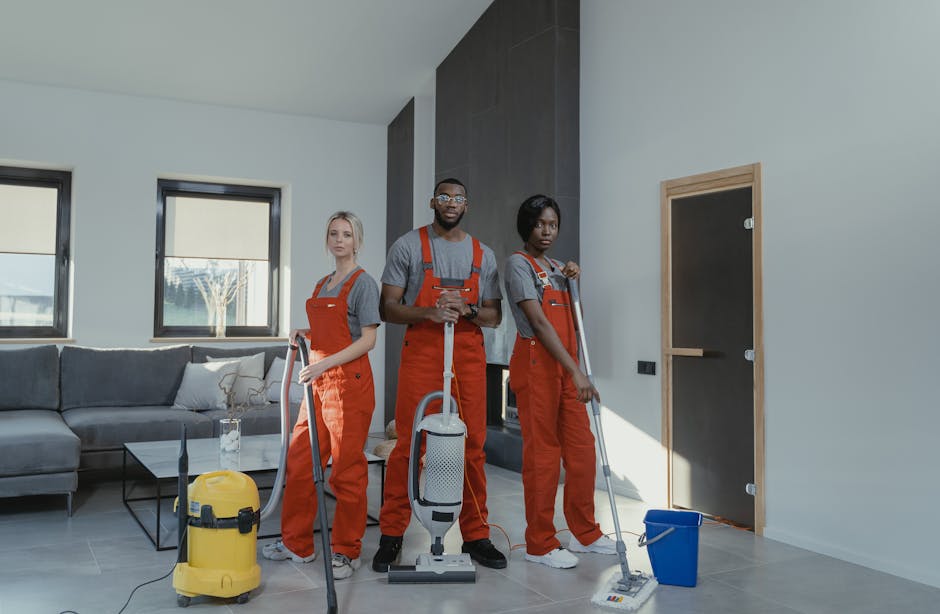 Three professional cleaners from Knightsbridge Cleaner, dressed in grey shirts and orange overalls, are performing surface cleaning in a modern living room. They are equipped with vacuum cleaners and